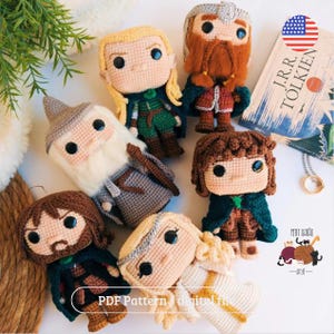 Pode incluir: Uma coleção de bonecas de crochê feitas à mão inspiradas nos personagens de "O Senhor dos Anéis" de J.R.R. Tolkien. As bonecas apresentam roupas e traços faciais detalhados, com um livro e um anel no fundo. As bonecas são de várias cores.