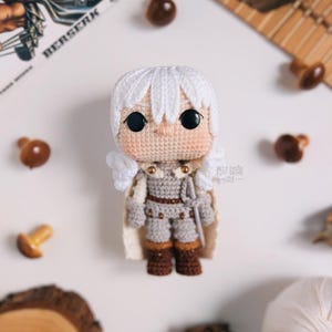 May include: A handmade crochet doll of a character with white hair, wearing a gray outfit with brown boots and a white cape. The doll has large black eyes and is surrounded by decorative wooden mushrooms and a book with the title "BERSERK".