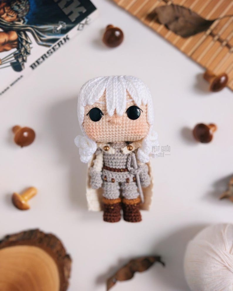 May include: A handmade crochet doll of a character with white hair, wearing a gray outfit with brown boots and a white cape. The doll has large black eyes and is surrounded by decorative wooden mushrooms and a book with the title "BERSERK".
