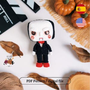 Pode incluir: Boneca de crochê feita à mão de um personagem da série de filmes de terror Saw. A boneca tem um rosto branco com cabelo preto, bochechas vermelhas e uma gravata borboleta vermelha. Ela está vestida com um terno preto e sapatos vermelhos. A imagem inclui abóboras e folhas de outono.