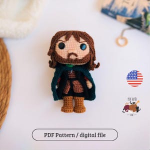 Pode incluir: Boneca de crochê feita à mão de um personagem com cabelo castanho, barba e capa verde. A boneca está vestida com uma roupa marrom e botas bege. O texto "PDF Pattern / digital file" está abaixo. Uma pequena bandeira americana está no fundo.