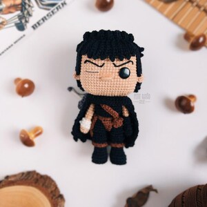 May include: Crocheted doll of a character with black hair, a black cape, and brown accents. The doll has a winking expression and is set against a white background with decorative elements. The word "BERSERK" is visible in the background.