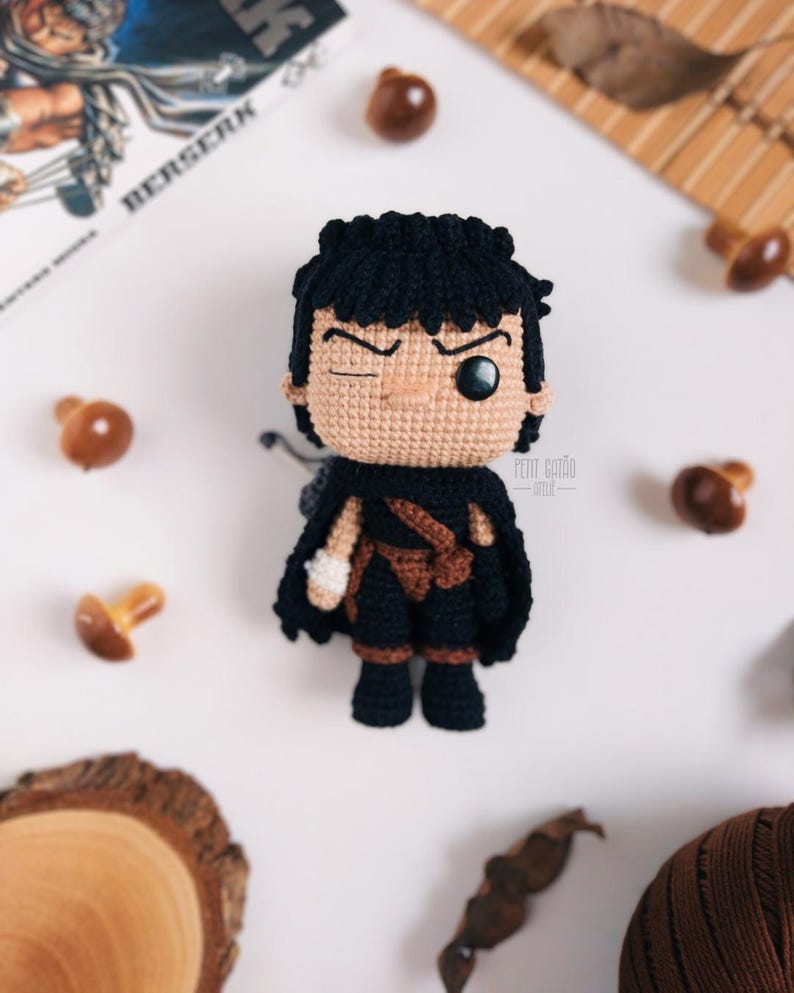 May include: Crocheted doll of a character with black hair, a black cape, and brown accents. The doll has a winking expression and is set against a white background with decorative elements. The word "BERSERK" is visible in the background.