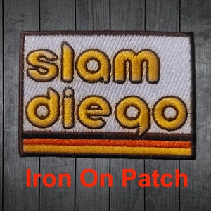 May include: Iron-on patch with the text "slam diego" in yellow outlined letters on a white background with a brown border and orange and yellow stripes.