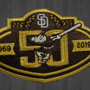 May include: A brown and gold embroidered patch with a baseball player swinging a bat. The patch has the text "1969" and "2019" on either side of the player and the letter "SD" in the top center.