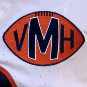 May include: Close-up of a white jersey with an orange and navy blue embroidered patch. The patch is in the shape of a football and features the letters "VMH". The jersey has small black dots.