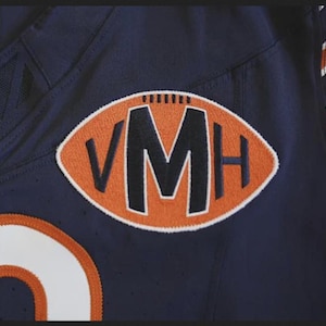 May include: Navy blue football jersey with an orange and white patch. The patch is in the shape of a football and features the letters "VMH" in black. The jersey has white and orange stripes on the shoulder.