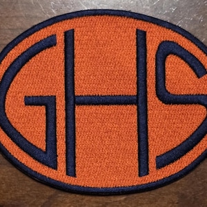 May include: An oval-shaped embroidered patch featuring the letters GHS in dark navy blue thread centered on a bright orange background. The patch has a pointed, stylized border in dark navy blue that outlines the entire shape of the design.