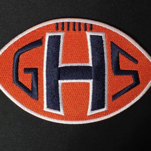 May include: An oval-shaped embroidered patch designed to resemble a football. The patch features a bright orange background with a white border and dark navy blue letters GHS centered in the middle, along with dark navy blue stitching for laces.