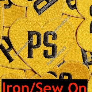 May include: A close-up of a pile of yellow heart-shaped iron-on patches with the letter "P" and "S" in black. The text "Iron/Sew On" is printed in red at the bottom of the image.