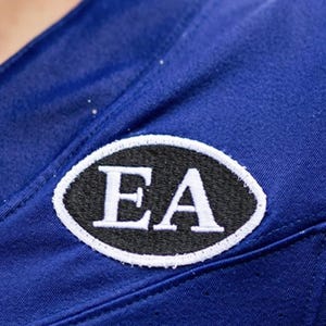 May include: A vibrant royal blue garment featuring an oval-shaped embroidered patch. The patch has a black background with the white capital letters EA stitched in the center, surrounded by a thick white border stitched onto the blue fabric.