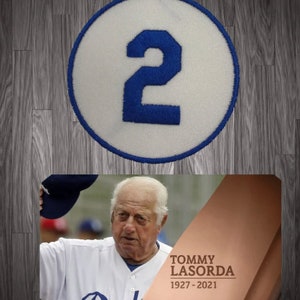 May include: A white circle with a blue number "2" on it. Below the circle is a photo of Tommy Lasorda in a Los Angeles Dodgers uniform with the text "TOMMY LASORDA 1927-2021".