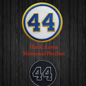 May include: Two embroidered patches featuring the number 44. The top patch is white with blue and yellow trim. The bottom patch is black with white stitching. The text "Hank Aaron Memorial Patches" is below the patches.