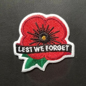 May include: Red embroidered poppy flower with a black center and green leaves. The poppy has a white border and a black ribbon with the text "Lest We Forget" in white.