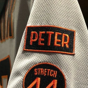 May include: A white baseball jersey with orange trim. The jersey has the word "PANIK" on the front and the number "44" on the sleeve. The sleeve also has a black patch with the word "PETER" in orange lettering. The patch is surrounded by an orange border.
