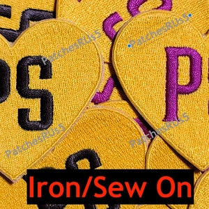May include: A stack of gold heart-shaped iron-on or sew-on patches with the letters "PS" in black and purple. The text "Iron/Sew On" is printed below the patches.