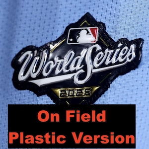 May include: A black, white, and gold World Series 2025 patch. The patch features the MLB logo and the words "World Series" in white lettering. The patch is on a light blue fabric. The text "On Field Plastic Version" is in a black rectangle.