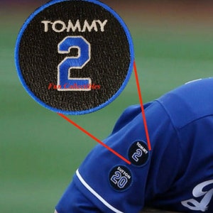 May include: A black and blue embroidered patch with the name "TOMMY" and the number "2" in white. The patch is sewn onto a blue baseball jersey.