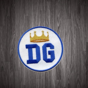 May include: A white circular patch with a blue embroidered "DG" and a gold crown above the letters. The patch is on a dark wood background.
