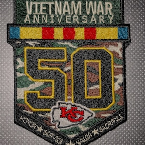 May include: A commemorative patch for the Vietnam War 50th Anniversary. The patch features the text "VIETNAM WAR ANNIVERSARY" above a shield-shaped design with the number "50" in black over a camouflage pattern. The Kansas City Chiefs logo is also present.