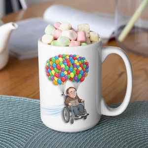 May include: A white ceramic mug filled with pastel-colored marshmallows. The front of the mug features a cartoon illustration of a person in a wheelchair being lifted into the air by a large cluster of colorful balloons, with a light blue wind trail.