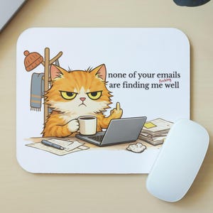 May include: White mousepad with a cartoon illustration of an orange cat giving the middle finger while working on a laptop. The text on the mousepad reads "none of your emails are finding me well."
