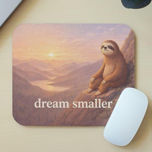 May include: A square mousepad featuring a sloth sitting on a mountain, with a sunset landscape in the background. The text "dream smaller" is printed on the mousepad. A white mouse and pencils are also visible.