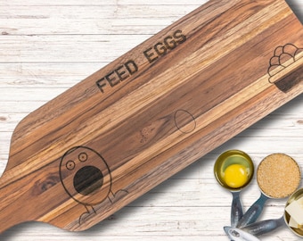以下が含まれることがあります： 「FEED EGGS」という文字が刻まれた木製のまな板。まな板には、目が3つで口を開けた漫画の鶏が刻まれています。また、卵の入った籠も刻まれています。