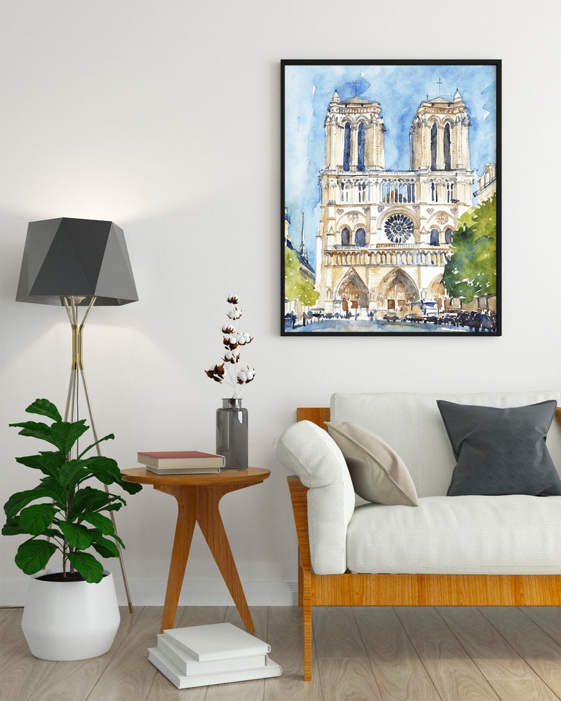 Op de afbeelding: Een aquarel schilderij van de Notre Dame kathedraal in Parijs, Frankrijk. Het schilderij is zwart ingelijst en hangt aan een witte muur boven een witte bank met een houten frame. Een kleine houten bijzettafel met een boek erop staat voor de bank.