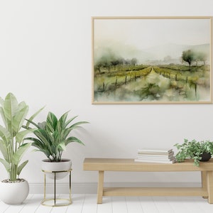 Op de afbeelding: Een ingelijst aquarel schilderij van een mistig landschap met een pad dat door een veld met groene bomen en struiken loopt. Het schilderij hangt aan een witte muur boven een houten bank met een stapel boeken en twee planten in potten.