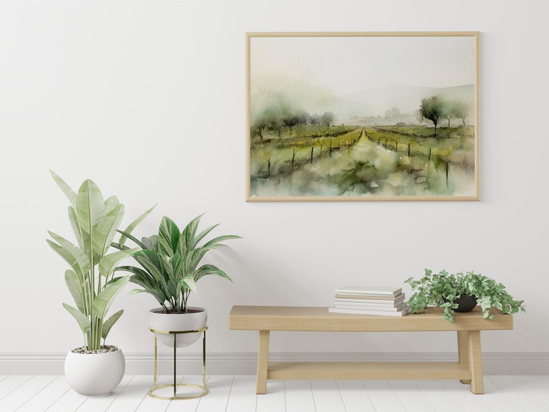 Op de afbeelding: Een ingelijst aquarel schilderij van een mistig landschap met een pad dat door een veld met groene bomen en struiken loopt. Het schilderij hangt aan een witte muur boven een houten bank met een stapel boeken en twee planten in potten.