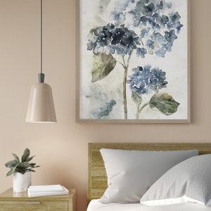 May include: A watercolor painting of blue hydrangeas with green leaves on a white background. The painting is framed in a light wood frame and is hanging on a light beige wall.
