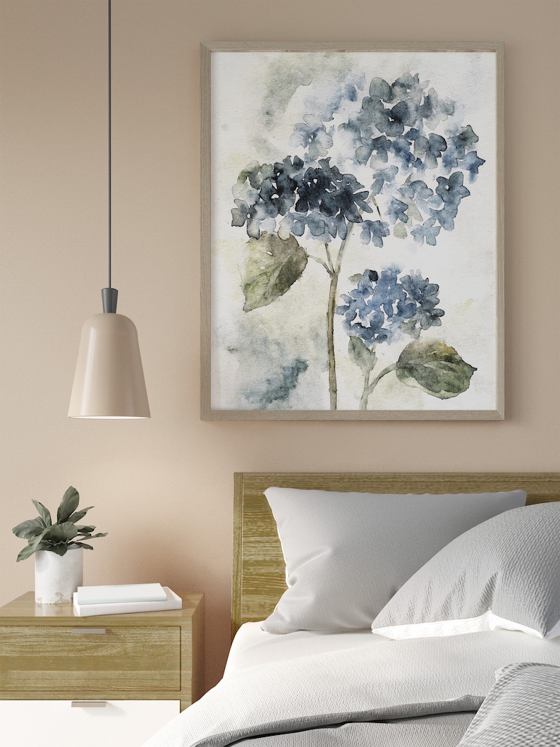 May include: A watercolor painting of blue hydrangeas with green leaves on a white background. The painting is framed in a light wood frame and is hanging on a light beige wall.