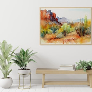 Puede incluir: Una pintura de acuarela de un paisaje des&eacute;rtico con formaciones rocosas rojas, arbustos verdes y un cielo azul. La pintura est&aacute; enmarcada en un marco de madera clara y est&aacute; colgada en una pared blanca. Hay un banco de madera con una pila de libros y dos plantas en macetas delante de la pintura.
