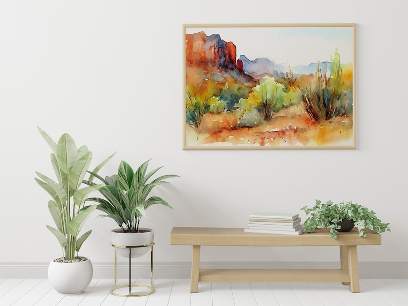 Puede incluir: Una pintura de acuarela de un paisaje des&eacute;rtico con formaciones rocosas rojas, arbustos verdes y un cielo azul. La pintura est&aacute; enmarcada en un marco de madera clara y est&aacute; colgada en una pared blanca. Hay un banco de madera con una pila de libros y dos plantas en macetas delante de la pintura.