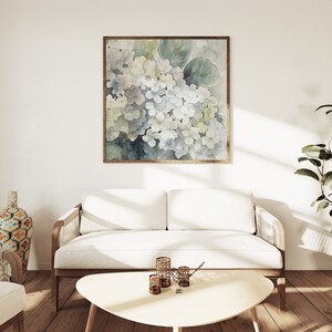 May include: A square framed watercolor painting of white and blue hydrangeas hangs above a white sofa. The painting has a gold frame. The room has a modern design with a wooden coffee table, a decorative vase, and a potted plant.