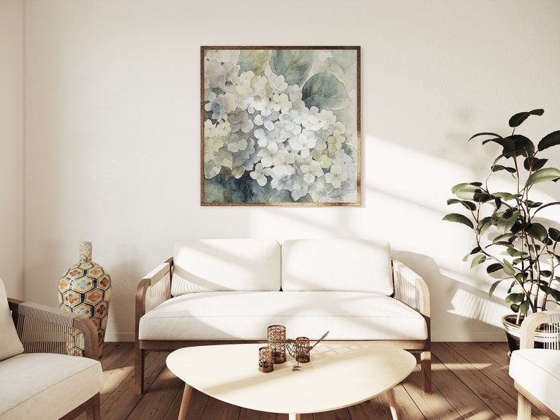 May include: A square framed watercolor painting of white and blue hydrangeas hangs above a white sofa. The painting has a gold frame. The room has a modern design with a wooden coffee table, a decorative vase, and a potted plant.