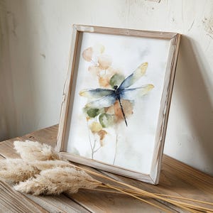 May include: Watercolor print of a dragonfly with blue and yellow wings, set against a backdrop of muted green and orange foliage. The artwork is framed in a rustic, light-colored wooden frame, creating a natural aesthetic. The artwork is displayed on a wooden surface.