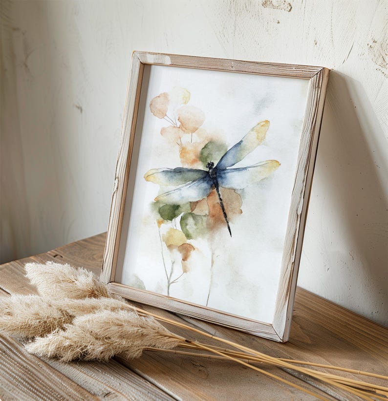 May include: Watercolor print of a dragonfly with blue and yellow wings, set against a backdrop of muted green and orange foliage. The artwork is framed in a rustic, light-colored wooden frame, creating a natural aesthetic. The artwork is displayed on a wooden surface.