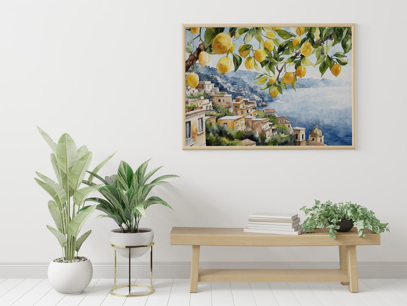 May include: A watercolor painting of a lemon tree with yellow lemons hanging from the branches. The tree is in front of a cityscape with buildings on a hillside overlooking the ocean. The painting is framed in a light wood frame and hanging on a white wall.