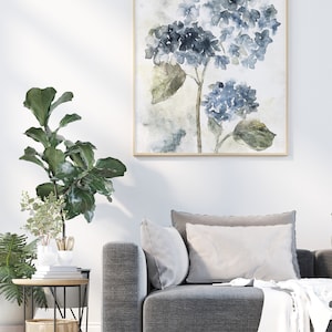 May include: A watercolor painting of blue hydrangeas with green leaves in a light gray frame. The painting is hanging on a white wall above a gray couch with white pillows.