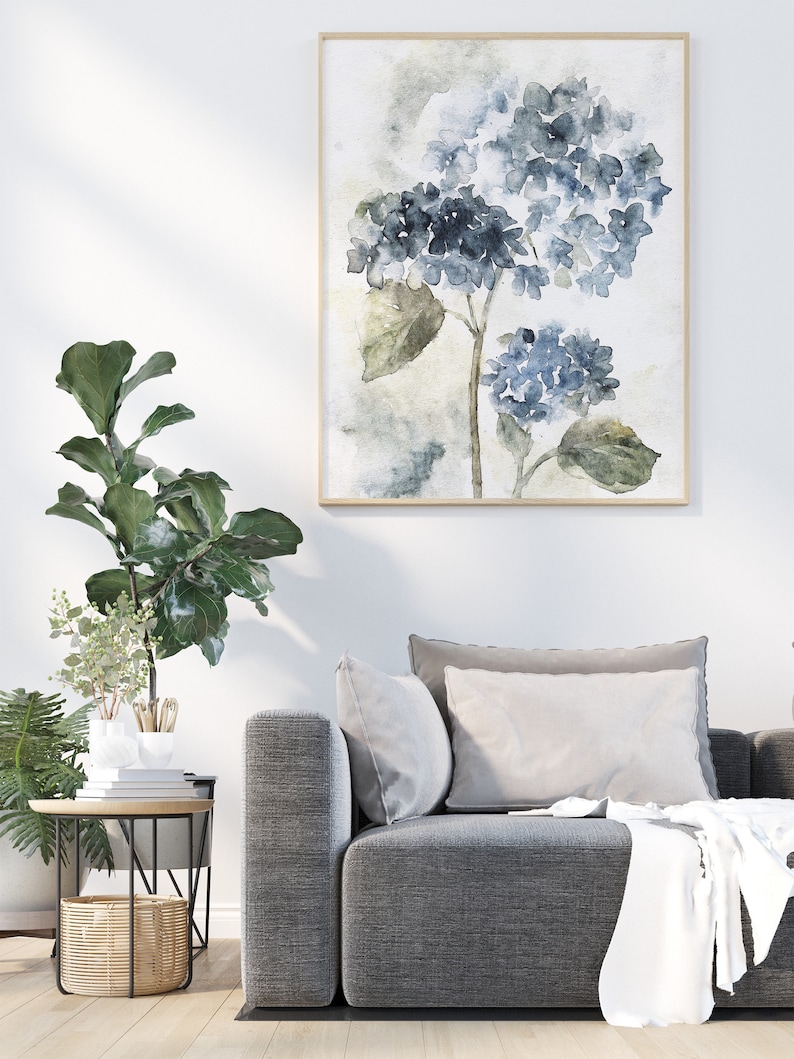May include: A watercolor painting of blue hydrangeas with green leaves in a light gray frame. The painting is hanging on a white wall above a gray couch with white pillows.