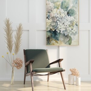 May include: A watercolor painting of white and blue hydrangeas in a wooden frame hangs above a green armchair with wooden arms and legs. Dried flowers and pampas grass are in vases, creating a serene, neutral-toned interior design.