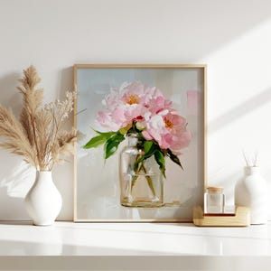 May include: A framed painting of pink peonies in a clear glass vase. The artwork has a light background and a natural wood frame. A white vase with dried pampas grass and a small glass jar with a wooden base are also visible.