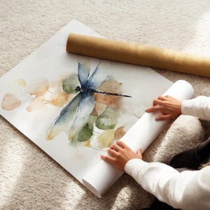 May include: A watercolor print of a dragonfly with blue, green, and brown hues. The artwork is on a white sheet of paper, with a rolled-up white paper tube and a brown cardboard tube nearby. The image is on a beige carpet.