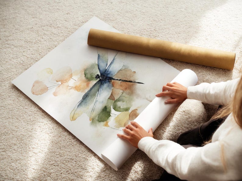 May include: A watercolor print of a dragonfly with blue, green, and brown hues. The artwork is on a white sheet of paper, with a rolled-up white paper tube and a brown cardboard tube nearby. The image is on a beige carpet.