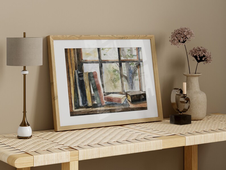 Puede incluir: Una pintura al agua enmarcada de una ventana con libros apilados en un alf&eacute;izar. La pintura est&aacute; en un marco de madera clara y est&aacute; colgada en una pared sobre una mesa de madera clara con una parte superior tejida.