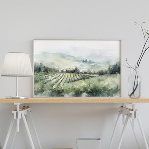 May include: A watercolor painting of a vineyard landscape with rolling hills and a foggy sky. The painting is framed in a simple white frame and is displayed on a wooden table with a white lamp and a vase with branches.