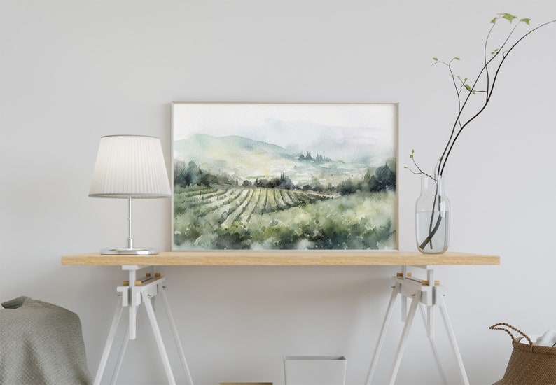 May include: A watercolor painting of a vineyard landscape with rolling hills and a foggy sky. The painting is framed in a simple white frame and is displayed on a wooden table with a white lamp and a vase with branches.