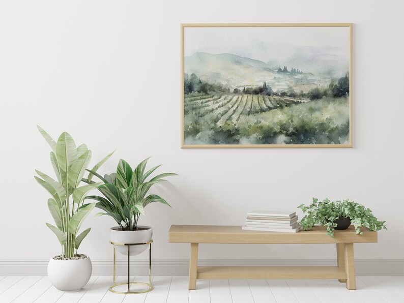 May include: A framed watercolor painting of a vineyard landscape with rolling hills and a hazy sky. The painting is hanging on a white wall above a wooden bench with a stack of books and two potted plants.